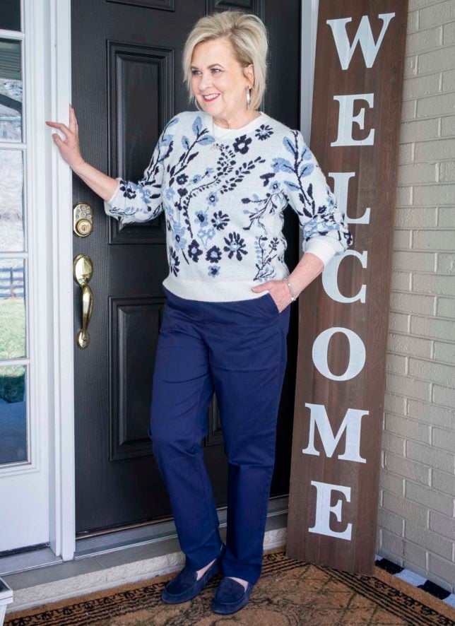 Fashion Blogger 50 Is Not Old is wearing a white sweater with a blue floral pattern, navy chino pants, and navy loafers