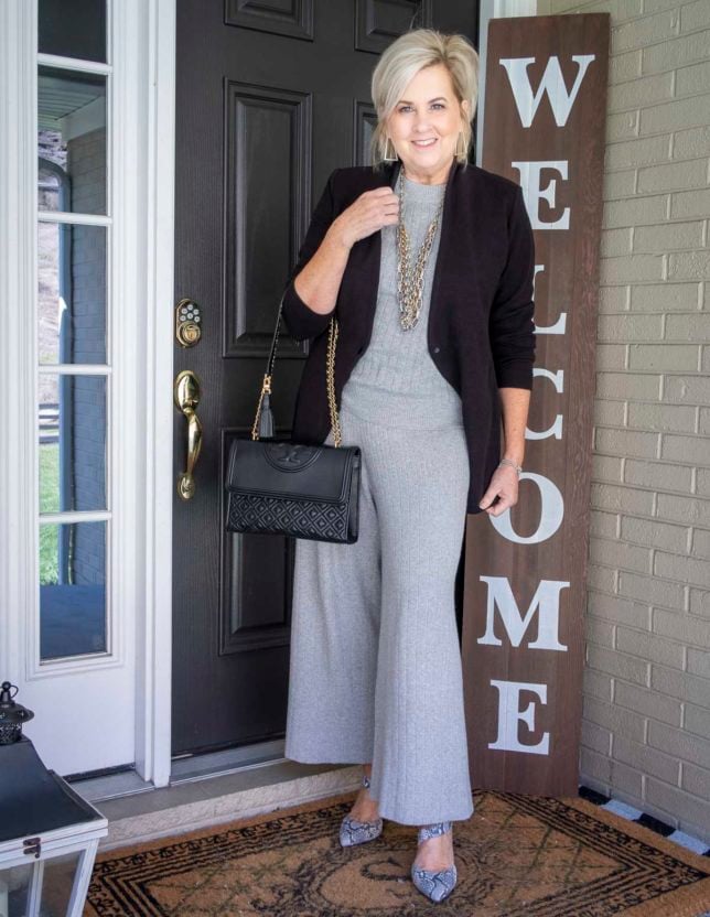 Fashion Blogger 50 Is Not Old is wearing a gray cropped loungewear set with a black blazer and gold jewelry, a Tory Burch handbag, and snakeskin heels