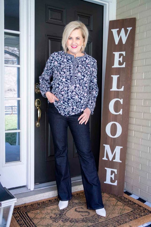 Fashion Blogger 50 Is Not Old is wearing a floral tunic, dark wash flare jeans, and white ankle boots