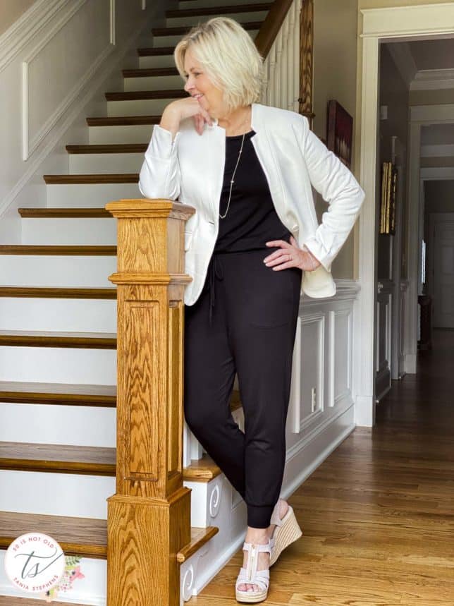 Fashion Blogger 50 Is Not Old is wearing a black jumpsuit and a white notched collar blazer from Gibsonlook and leaning on a stairpost