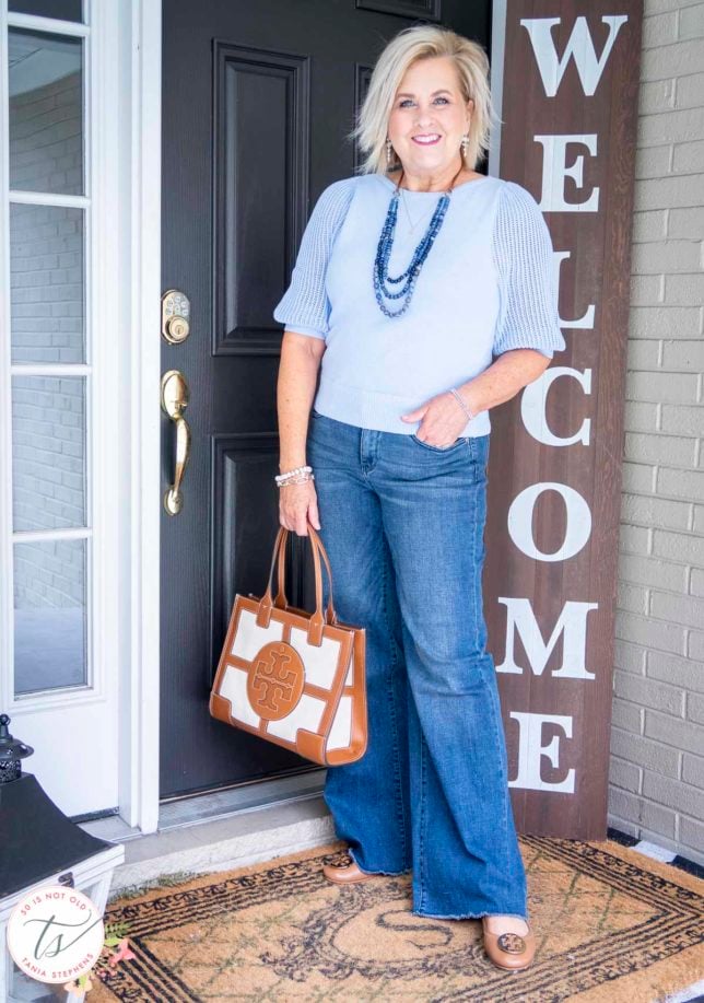 Fashion Blogger 50 Is Not Old is standing with her hand in her pocket in a pair of wide-leg jeans with a light blue sweater, and a Tory Burch tote