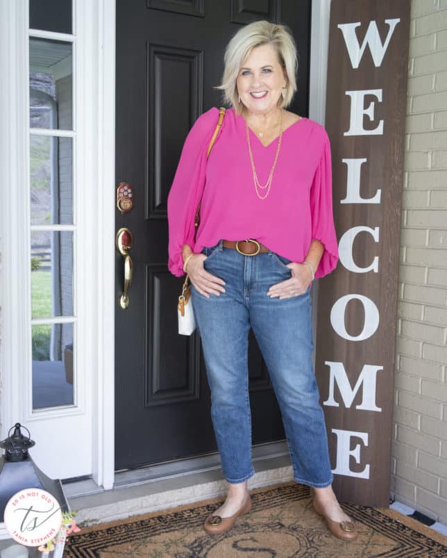Fashion Blogger 50 Is Not Old is wearing a v-neck pink blouse, a pair of straight leg jeans, ballet flats, and a Dooney & Bourke handbag