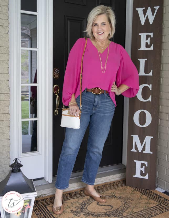 Fashion Blogger 50 Is Not Old is wearing a v-neck bright pink blouse, a pair of straight leg jeans, ballet flats, and a Dooney & Bourke handbag