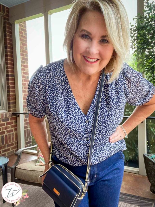 Fashion Blogger 50 Is Not Old is standing in a sunporch showing a blue and white print blouse and a navy crossbody bag