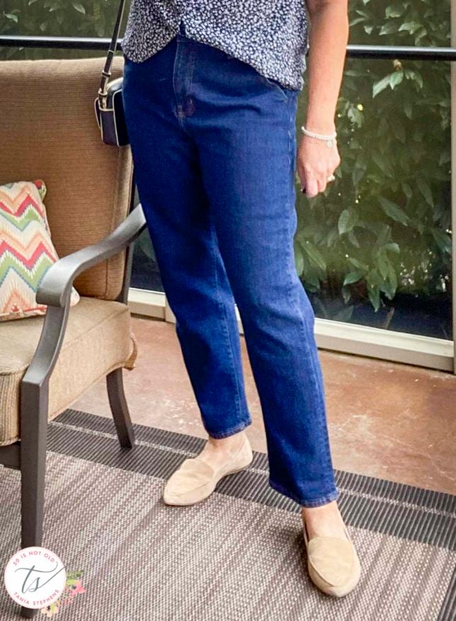 Fashion Blogger 50 Is Not Old is standing in a sunporch wearing a pair of dark wash jeans and neutral flats