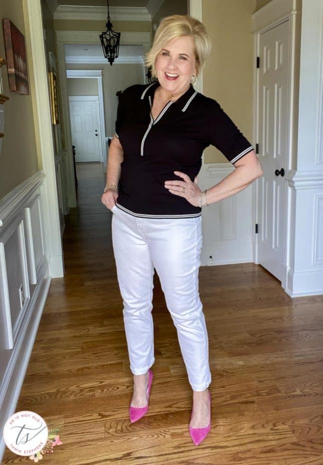 Fashion Blogger 50 Is Not Old is wearing a black polo sweater with white stripes on the edges, white straight-leg jeans, and bright pink suede pumps