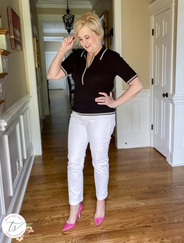 Fashion Blogger 50 Is Not Old is wearing a black polo sweater with white edging, white straight-leg jeans, and bright pink pumps