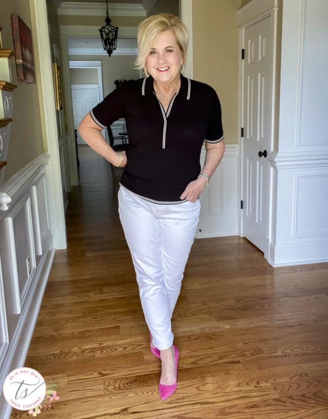 Fashion Blogger 50 Is Not Old is wearing a black polo sweater with white edging, white straight-leg jeans, and pink suede pumps