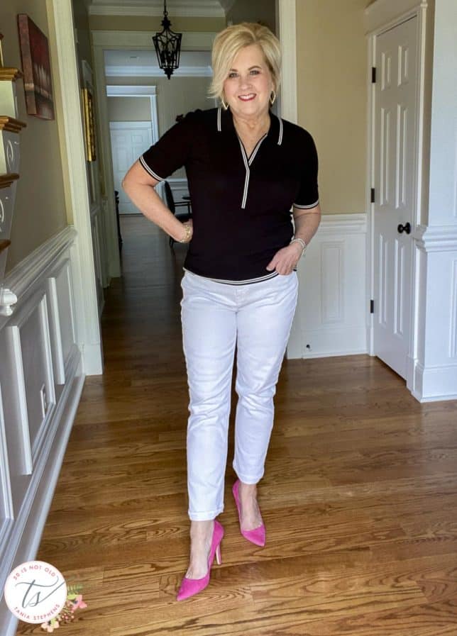 Fashion Blogger 50 Is Not Old is wearing a black polo sweater with white edging, white straight-leg jeans, and bright pink suede pumps