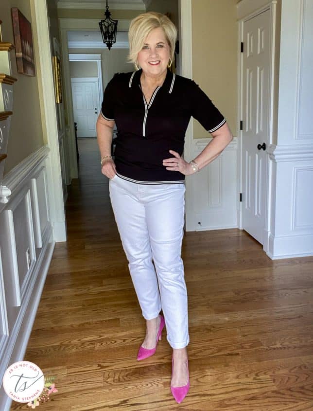 Fashion Blogger 50 Is Not Old is wearing a black polo sweater with white edging, white jeans, and bright pink suede pumps