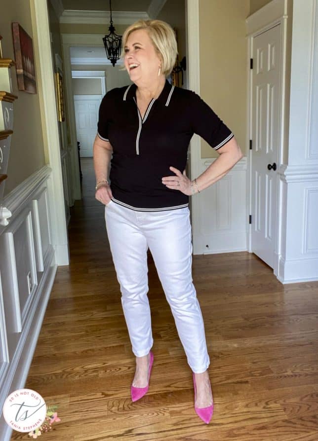Fashion Blogger 50 Is Not Old is laughing while wearing a black polo sweater with white edging, white straight-leg jeans, and bright pink suede pumps