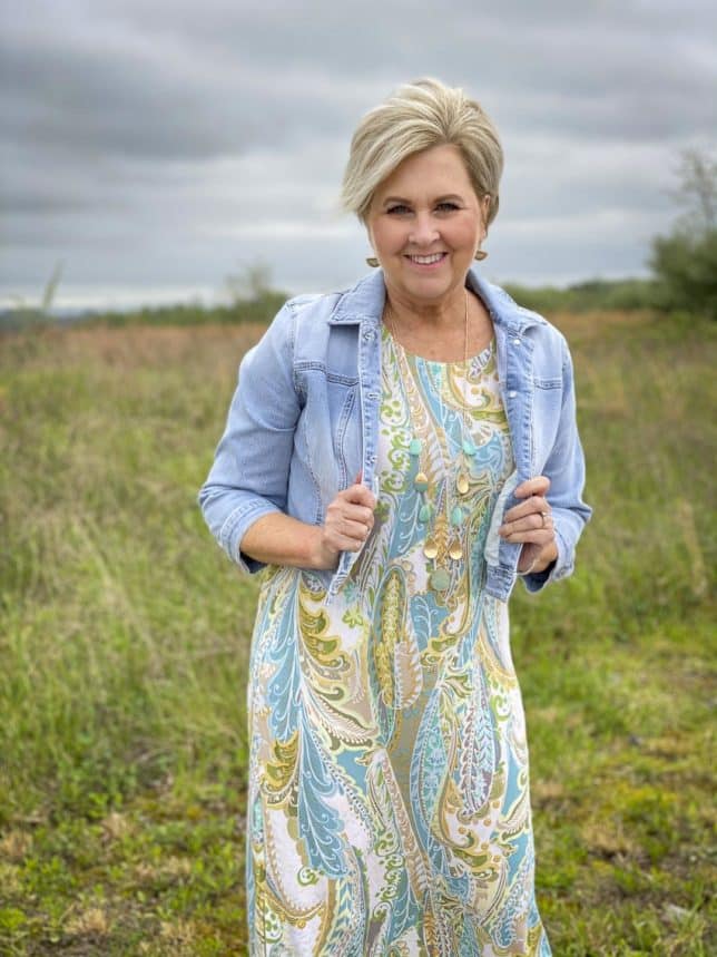 Fashion Blogger 50 Is Not Old is wearing a cropped denim jacket, a paisley print maxi dress, and a long necklace