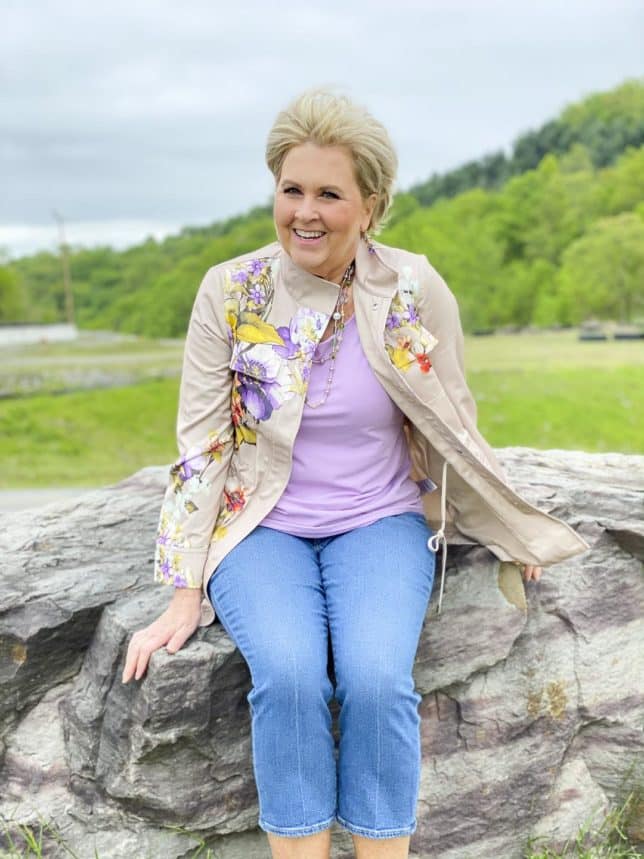 Fashion Blogger 50 Is Not Old is sitting on a large rock wearing a lavender tee with a floral satin utility jacket and cropped flare jeans from Chicos