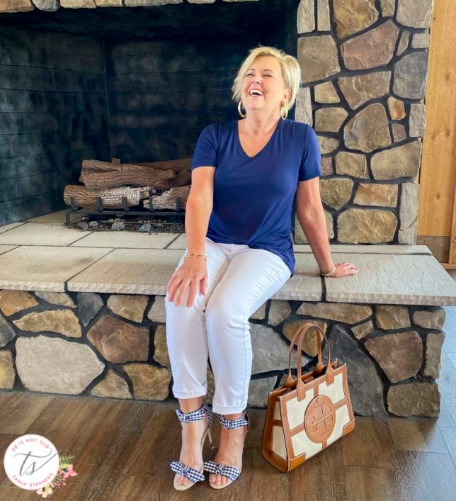 Fashion Blogger 50 Is Not Old is laughing hard while sitting on a fireplace wearing a navy top, white jeans, and gingham bow heels