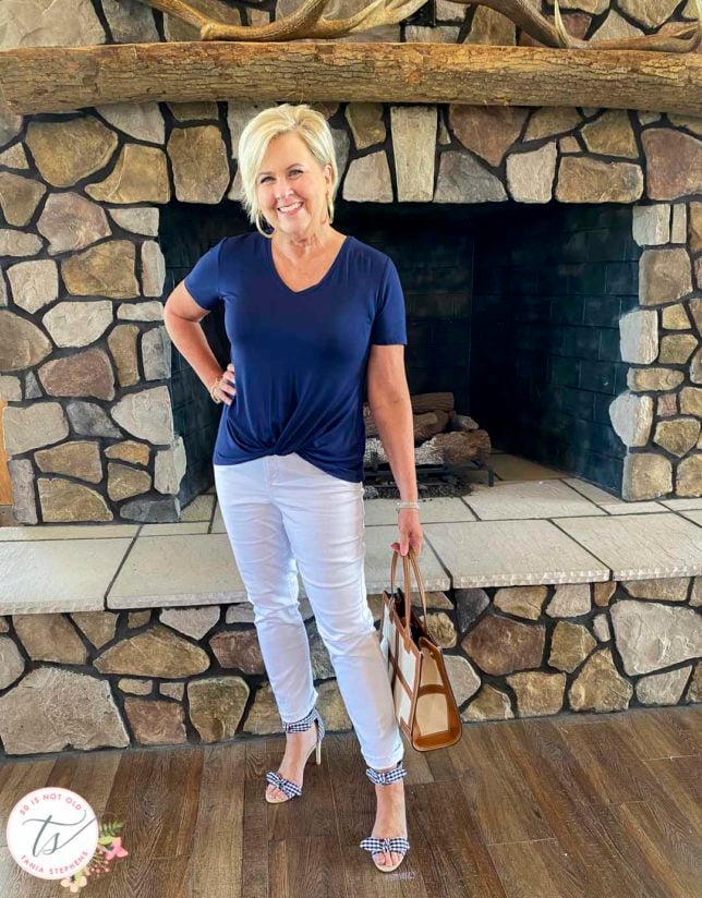 Fashion Blogger 50 Is Not Old is standing in front of a fireplace wearing a navy top, white jeans, and gingham bow heels