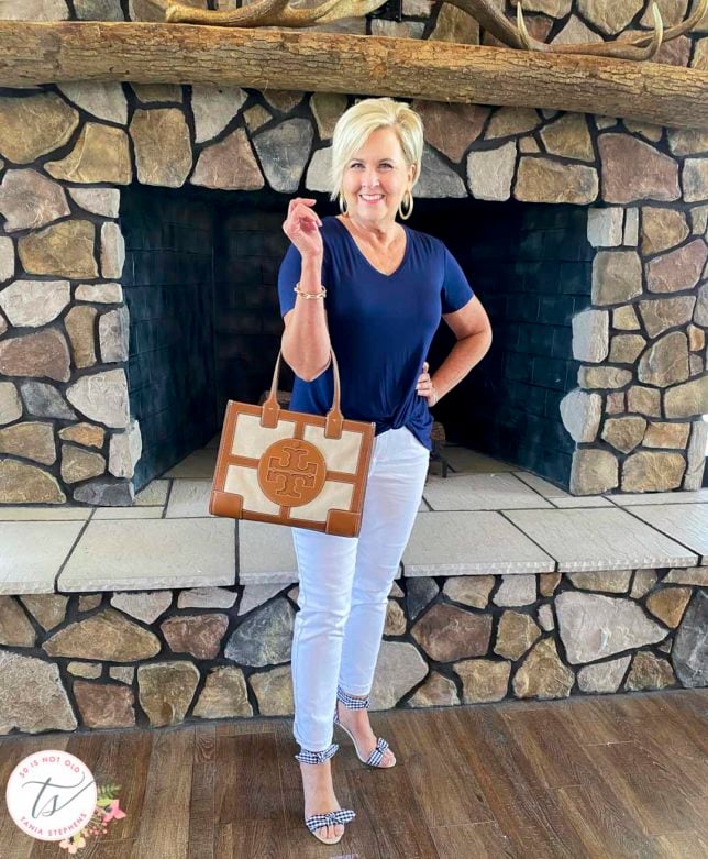 Fashion Blogger 50 Is Not Old is standing in front of a fireplace wearing a navy top, white jeans, carrying a Tory Burch handbag, and gingham bow heels