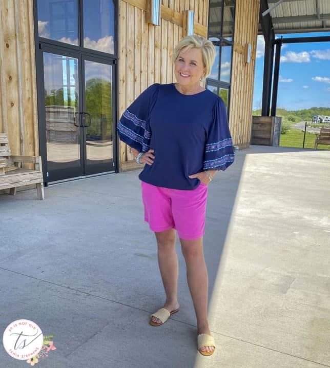 Fashion Blogger 50 Is Not Old is wearing a pair of bright pink colored shorts, a navy top with embroidered balloon sleeves, and raffia slide sandals
