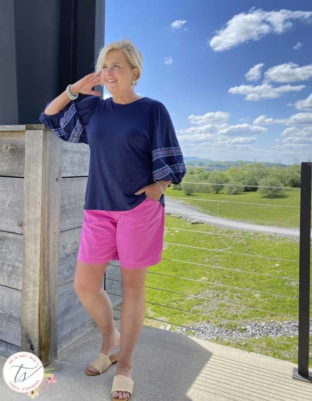 Fashion Blogger 50 Is Not Old is wearing a pair of bright pink shorts, a navy top with embroidered balloon sleeves, and raffia sandals
