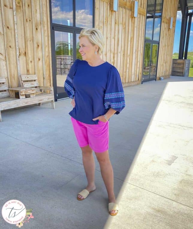Fashion Blogger 50 Is Not Old is looking to the side while wearing a pair of bright pink shorts, a navy top with embroidered balloon sleeves, and raffia slide sandals