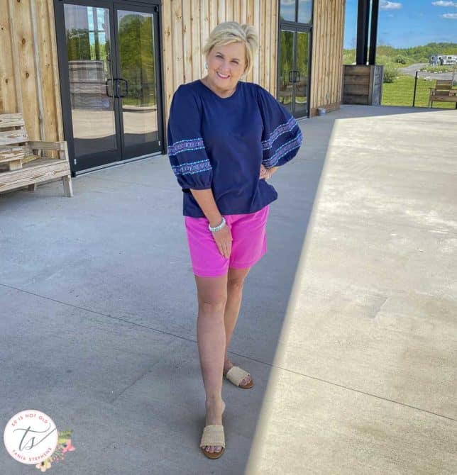 Fashion Blogger 50 Is Not Old is wearing a pair of bright pink shorts, a navy top with embroidered balloon sleeves, and slide sandals