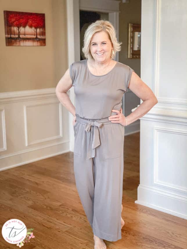 Fashion Blogger 50 Is Not Old is wearing a gray jumpsuit with an elastic waist and sash, and is barefooted