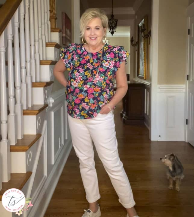 Fashion Blogger 50 Is Not Old is wearing a colorful floral top with flutter sleeves with a pair of white boyfriend jeans