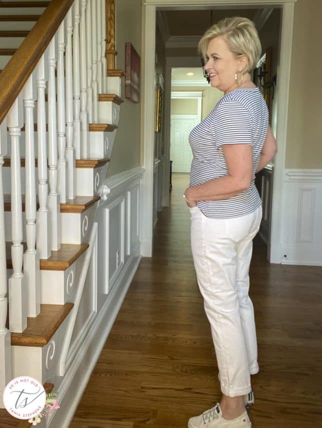 Fashion Blogger 50 Is Not Old is showing a side view of a blue and white striped tee with white boyfriend jeans and white sneakers