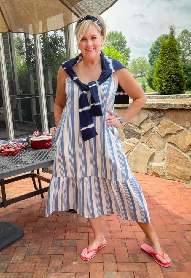 Fashion Blogger 50 Is Not Old is looking festive for the 4th of July in a blue and white striped sundress, a navy headband with stars, red flip-flops, and a navy tie-dyed sweater