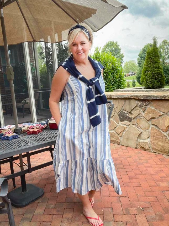 Fashion Blogger 50 Is Not Old is on a patio looking festive for the 4th of July in a blue and white striped sundress, a navy headband with stars, red flip-flops, and a navy tie-dyed sweater