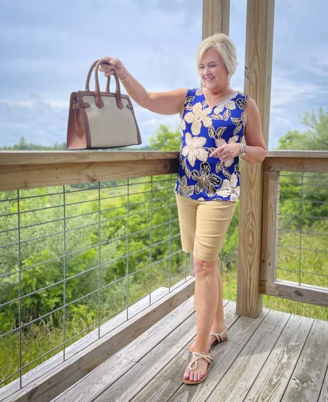 Fashion Blogger 50 Is Not Old is showing a canvas handbag while wearing a big, bold blue print sleeveless tank and 10" khaki shorts with gold braided sandals