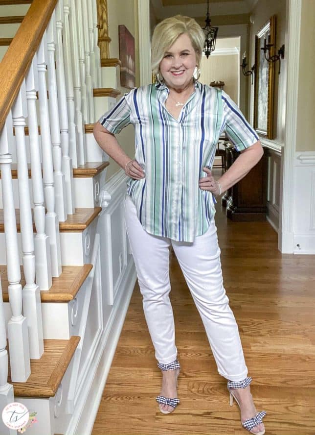 Fashion Blogger 50 Is Not Old is standing beside stairs and wearing a striped top with white jeans and navy and white gingham heels