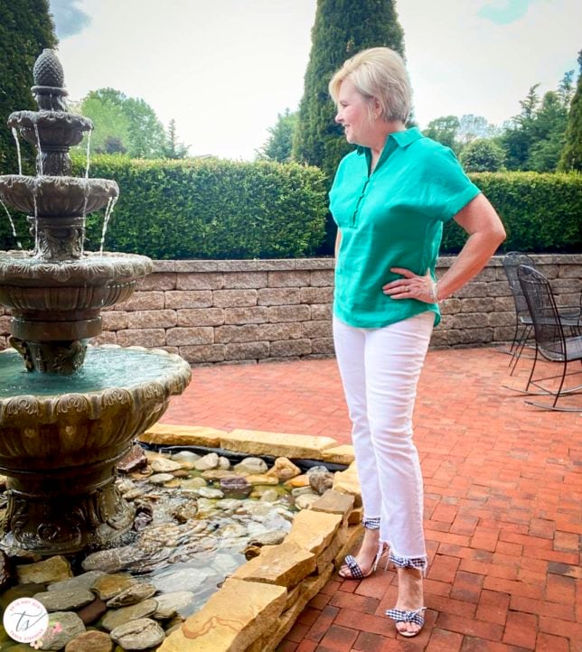 Fashion Blogger 50 Is Not Old is looking at a water fountain while wearing a jade linen pullover shirt with white crop jeans and navy and white gingham heels