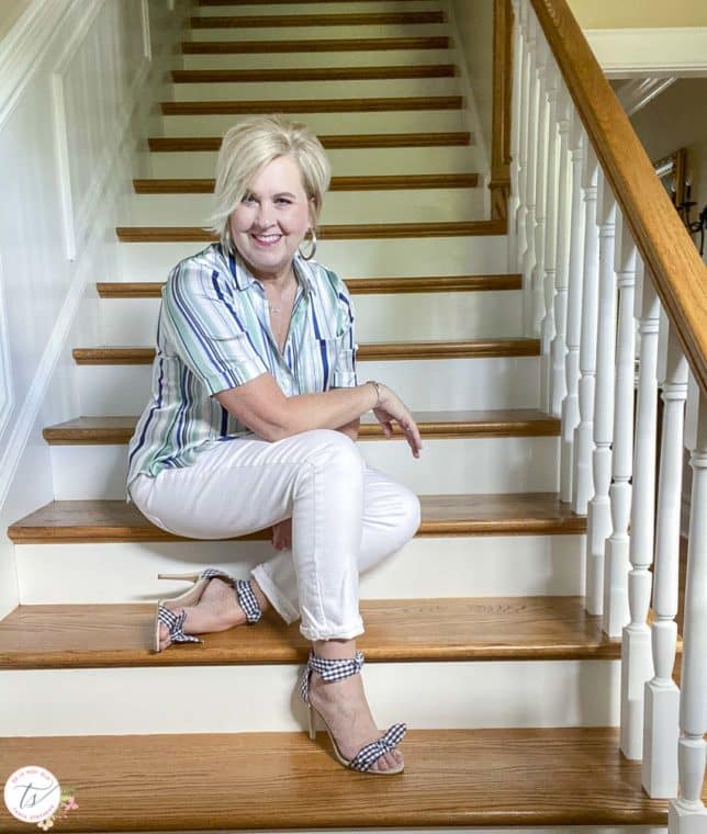 Fashion Blogger 50 Is Not Old is wearing a striped top with white jeans and navy and white gingham heels while sitting on stairs