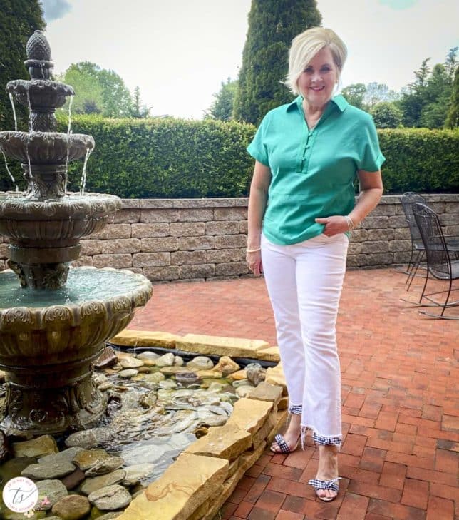 Fashion Blogger 50 Is Not Old is standing by a water fountain while wearing a jade linen pullover shirt with white crop jeans and navy and white gingham heels