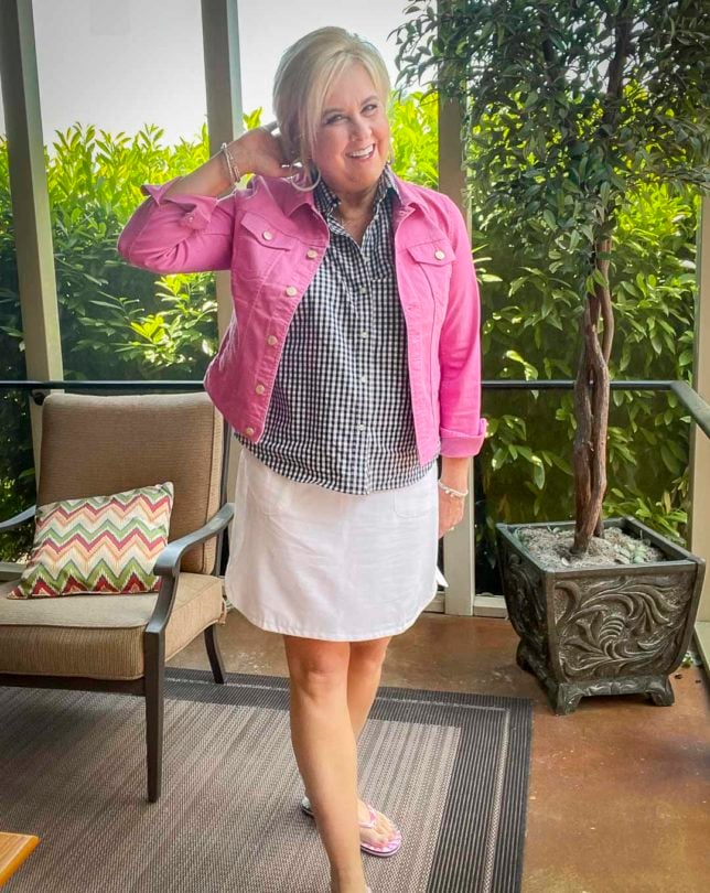 Fashion Blogger 50 Is Not Old is laughing while wearing an orchid jean jacket with a navy and white gingham button up sleeveless shirt with a white skort