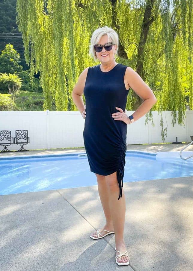 Fashion Blogger 50 Is Not Old is standing by a swimming pool wearing a black ruched dress with black sunglasses and white sandals