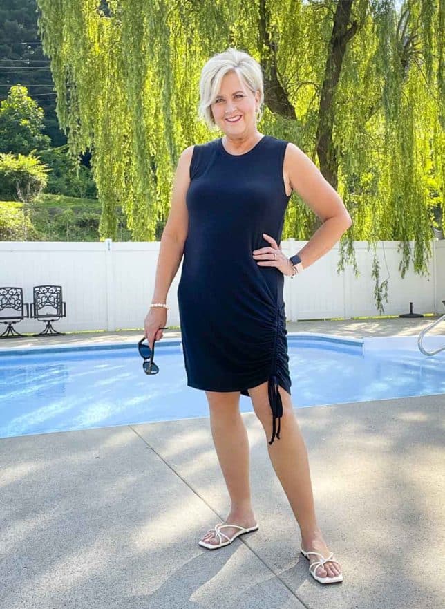 Fashion Blogger 50 Is Not Old is wearing a black ruched dress with black sunglasses in her hand and white sandals