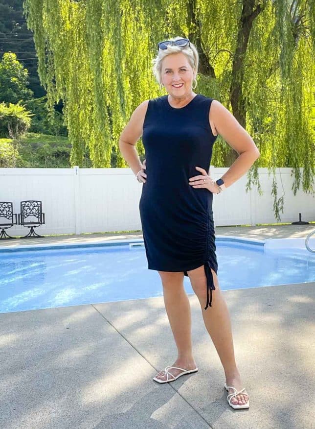 Fashion Blogger 50 Is Not Old is wearing a black ruched dress with black sunglasses on her head and white sandals