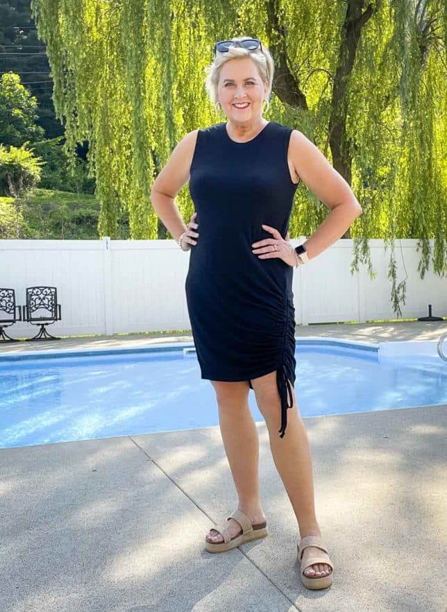 Fashion Blogger 50 Is Not Old is wearing a black ruched dress with black sunglasses on her head and platform sandals