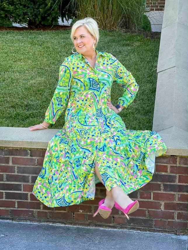 Fashion Blogger 50 Is Not Old is sitting on a brick wall in a colorful tropical midi shirt dress with bright pink suede pumps