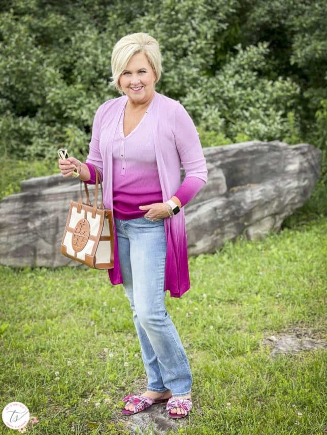 Fashion Blogger 50 Is Not Old is looking colorful in a pink ombre tank and matching coverup, bootcut jeans, floral slides, and is carrying a Tory Burch satchel