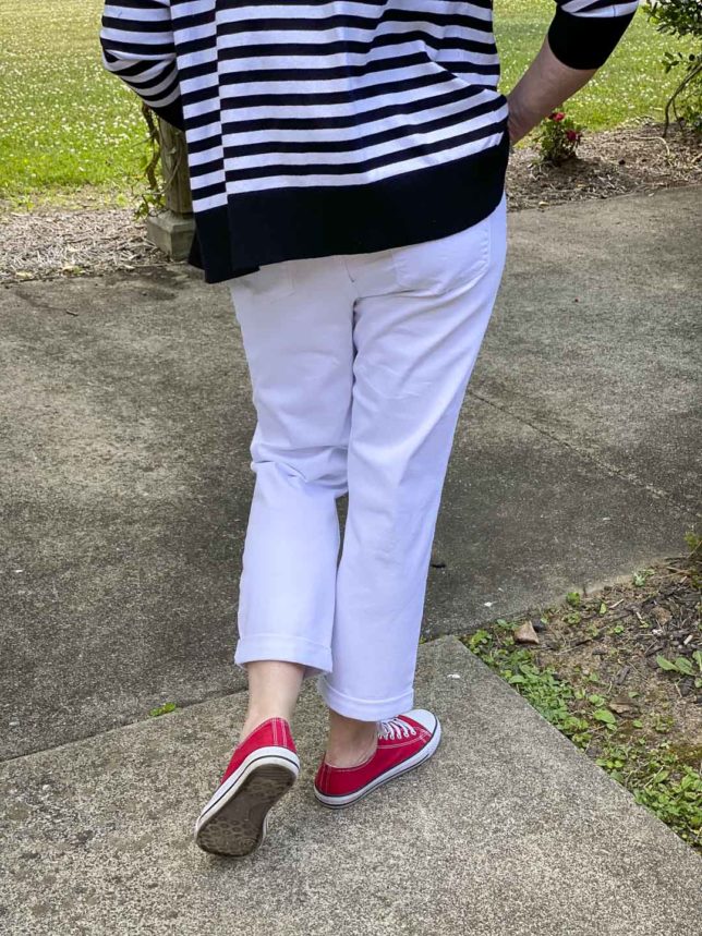Fashion Blogger 50 Is Not Old is showing the back of a black and white striped zip up sweater hoodie, white jeans, and red sneakers