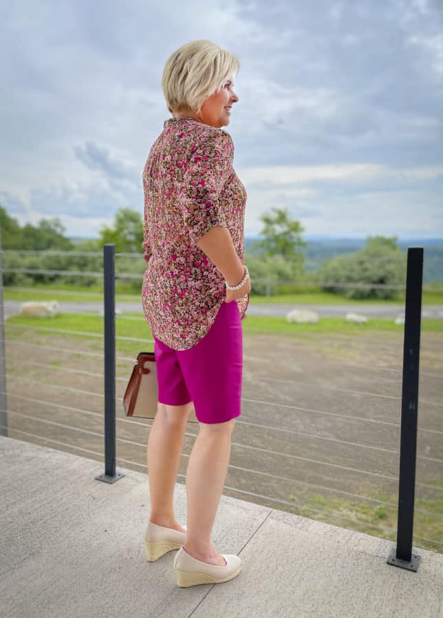 Fashion Blogger 50 Is Not Old is showing the back of a floral button-up blouse with orchid Bermuda shorts, and neutral wedge espadrilles