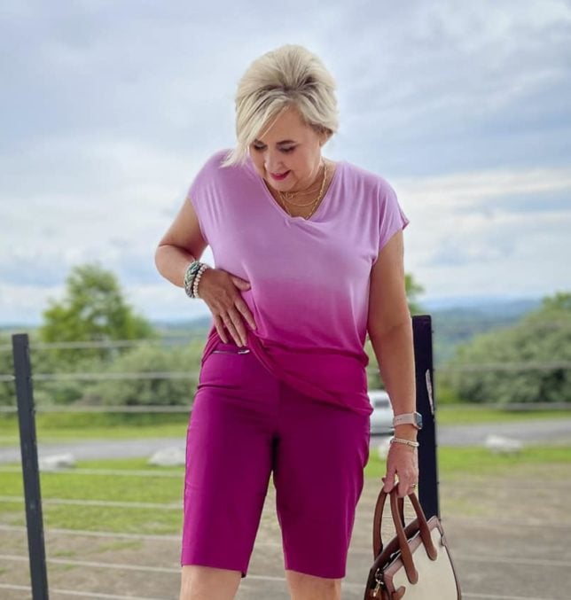 Fashion Blogger 50 Is Not Old is wearing an orchid ombre tee and showing the top of her orchid Bermuda shorts, Sketcher wedge sandals, and a canvas handbag