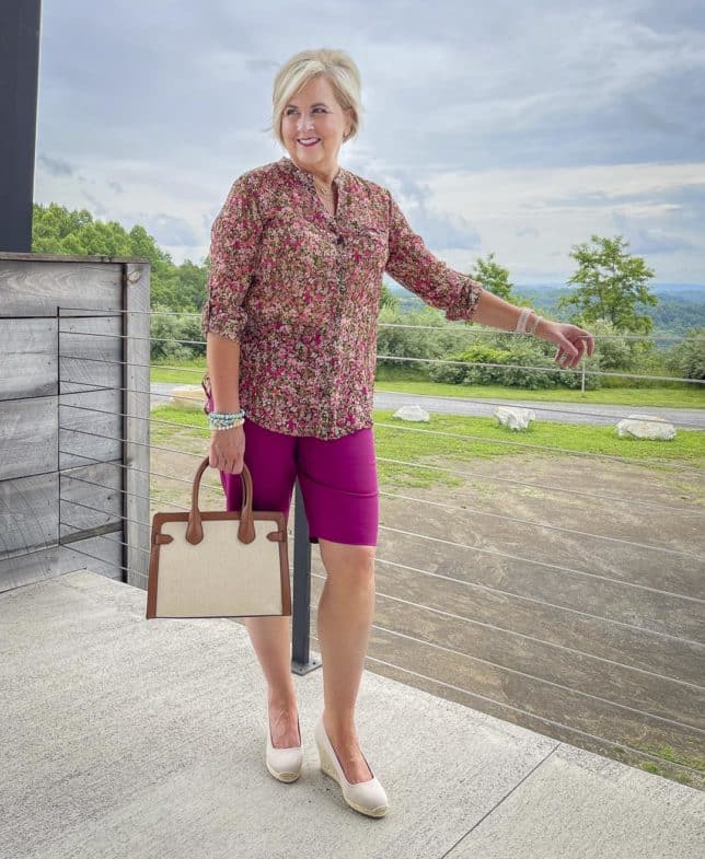 Fashion Blogger 50 Is Not Old is wearing a floral button-up blouse with orchid Bermuda shorts, neutral wedge espadrilles, and a canvas handbag