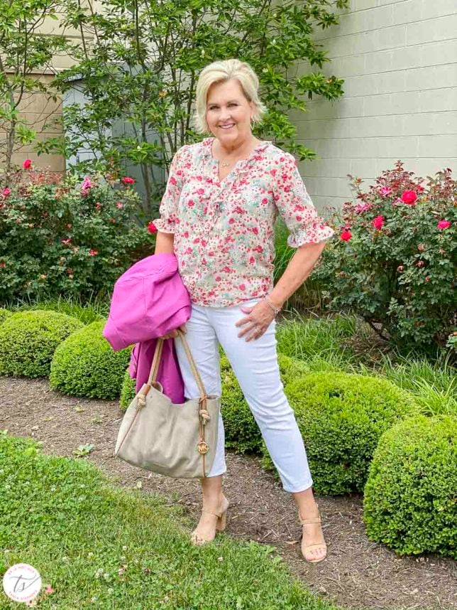 Fashion Blogger 50 Is Not Old is wearing a feminine floral blouse, white crop pants, cork heels, and carrying a orchid trench coat and a Michael Kors handbag