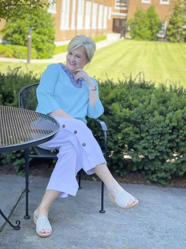 Fashion Blogger 50 Is Not Old is sitting at an outdoor table while wearing a beautiful aqua blue tunic with a patterned neckline and white side button crop pants with white slides