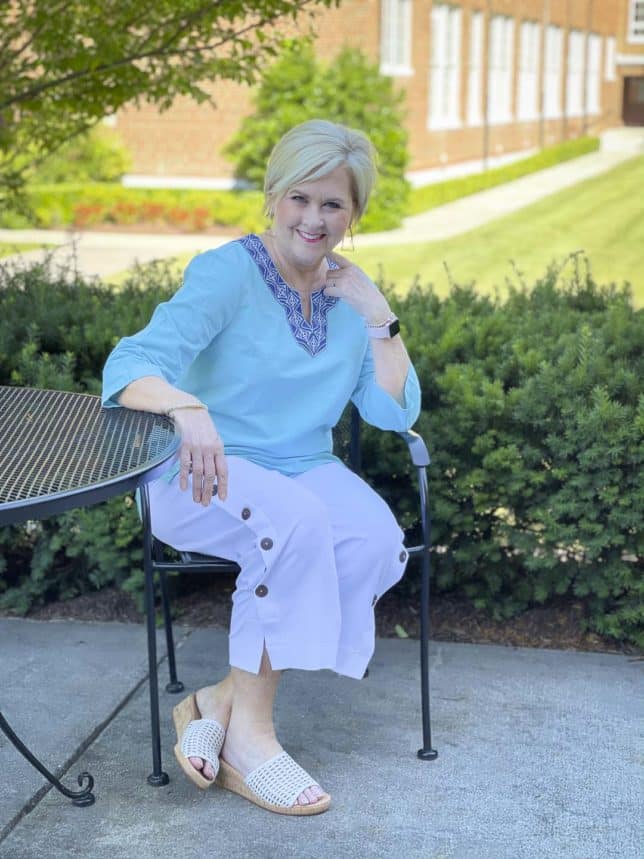 Fashion Blogger 50 Is Not Old is sitting at an outdoor table while wearing a beautiful aqua blue tunic and white side button crop pants