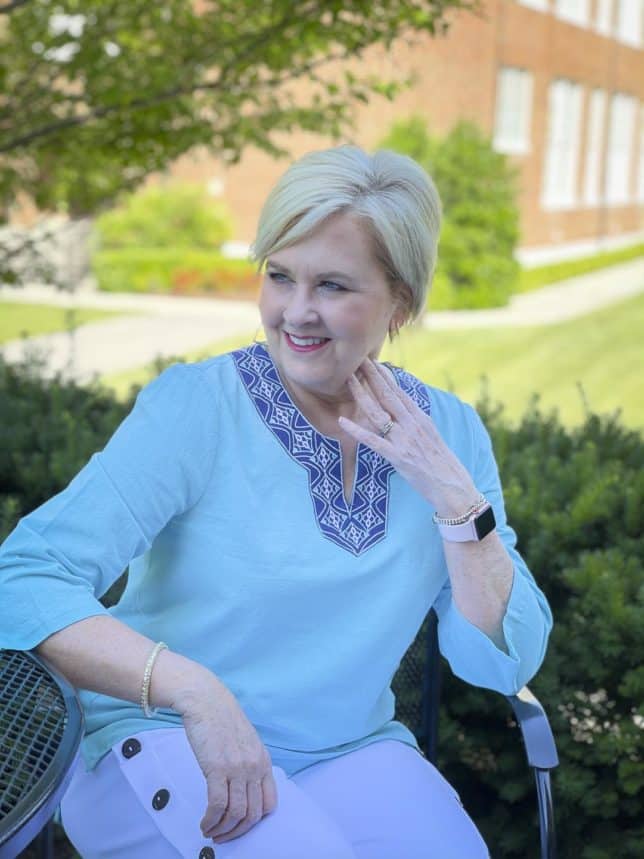 Fashion Blogger 50 Is Not Old is sitting at an outdoor table while wearing a beautiful aqua blue tunic with a unique neckline