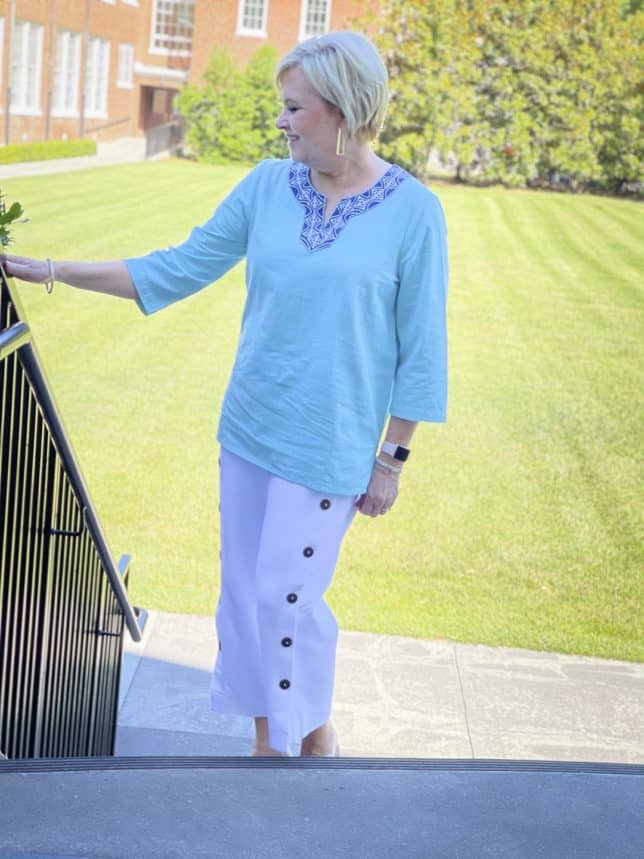 Fashion Blogger 50 Is Not Old is standing on stairs while wearing a beautiful aqua blue tunic with a patterned neckline and white side button crop pants